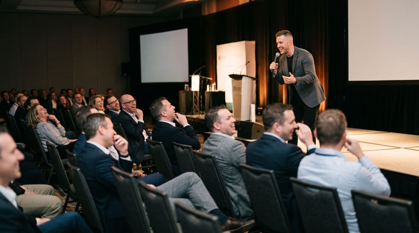 A comedian in a blazer performing on a conference stage while rows of professionals laugh